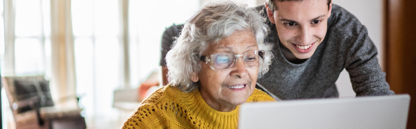 An older woman with glasses uses her laptop, helped by a younger man