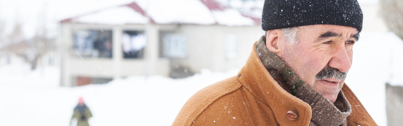 An older man with a moustache and a beanie hat, looking cold against a snowy background