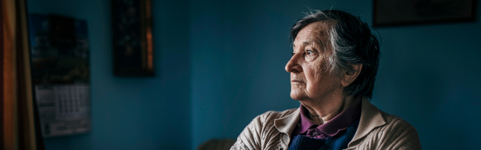 An older woman in a cardigan sits in a cold-looking dark room, looking out of the window